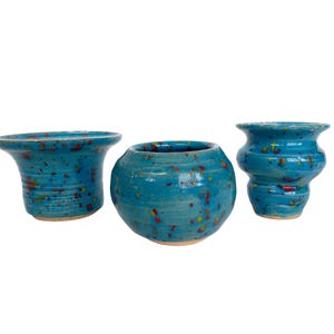 May include: Three handmade ceramic vases in varying shapes. The vases are a vibrant turquoise with speckled accents of red, yellow, and orange. The vases have a glossy finish and sit on a white background.
