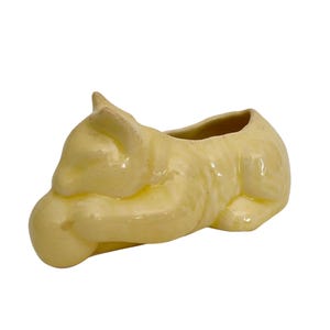 May include: A light yellow ceramic cat-shaped planter. The cat is curled up with its head resting on its paws. The planter has a glossy finish and a small opening at the top for plants or other items.