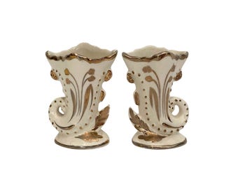 Könnte beinhalten: Zwei passende cremefarbene Keramikvasen mit goldenen Akzenten. Die Vasen haben eine hornartige Form mit ausgestellter Oberseite, einer dekorativen Spirale und goldenen floralen Designs. Die Ränder und Basen sind ebenfalls goldfarben.