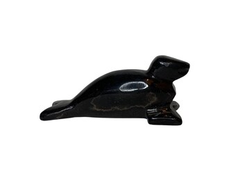 May include: A small, polished black seal figurine. The sculpture is smooth and glossy, with a rounded body and flipper details. The seal is lying down, with its head raised slightly.