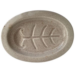 May include: Oval-shaped, speckled ceramic soap dish in a neutral beige color. The dish features a raised fishbone design in the center. The dish is designed to hold a bar of soap and has a slightly raised edge.
