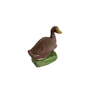 May include: A ceramic duck figurine with a brown body and a yellow beak. The duck is perched on a green base. The figurine has a textured surface and is set against a white background.