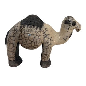 May include: A ceramic camel figurine featuring a cream-colored body with a dark brown crackle glaze pattern. The camel has a dark brown saddle area on its back, dark brown hooves, and dark brown facial markings around the eyes and nose.
