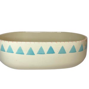 May include: Cream-colored ceramic bowl with a geometric design. The bowl has a rectangular shape with rounded edges. A row of blue triangles decorates the exterior, just below the rim. The rim is accented with a pink dotted line.