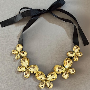 May include: A necklace featuring five butterfly-shaped clusters of faceted yellow crystals attached to a black grosgrain ribbon. The ribbon is tied in a bow at the top, and the crystal butterflies are arranged in a symmetrical arc along the band.