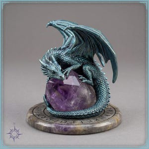 May include: A teal-colored dragon figurine with textured scales and spread wings, perched atop a large, faceted purple amethyst crystal. The entire sculpture rests on a circular, stone-like base featuring engraved runic symbols around the edge.