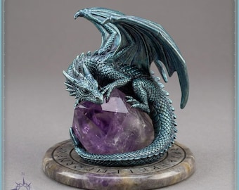 May include: A teal-colored dragon figurine with textured scales and spread wings, perched atop a large, faceted purple amethyst crystal. The entire sculpture rests on a circular, stone-like base featuring engraved runic symbols around the edge.