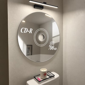 May include: A round wall mirror featuring a white graphic print of a CD-R disc with the text CD-R, Compact Disc Digital Audio, and 700MB. The mirror is mounted on a wall beneath a black horizontal light fixture, reflecting a room interior.