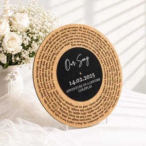 May include: A circular wooden plaque featuring a black center with the white text Our Song, 14.02.2025, and Adventure of a Lifetime Coldplay. The outer wooden border is engraved with song lyrics in a circular pattern around the black center.
