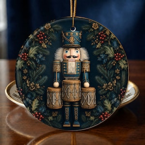 May include: A round ceramic ornament featuring a blue nutcracker soldier playing three drums. The nutcracker wears a matching blue uniform with gold accents and a tall hat. The background is a dark blue wreath of pine branches, holly leaves, and red berries.