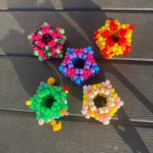 Pode incluir: Cinco pulseiras em forma de estrela feitas de contas de plástico coloridas. Uma é preta e verde neon, uma é vermelha, laranja e amarela, uma é roxa e azul, uma é verde brilhante com patinhos amarelos e uma é rosa pastel e amarela.