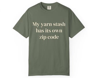 Puede incluir: Una camiseta de manga corta de color verde oliva con el texto "My yarn stash has its own zip code" impreso en color crema claro en el pecho. La prenda tiene un cuello redondo y un corte estándar, con una pequeña etiqueta en el cuello.