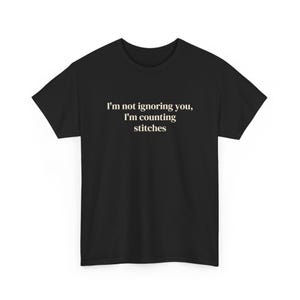May include: A black short-sleeved t-shirt featuring the white text "I'm not ignoring you, I'm counting stitches" printed in a clean, sans-serif font across the center of the chest area, set against a solid black fabric background.