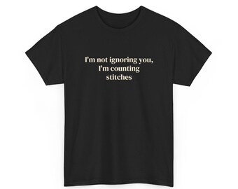 Puede incluir: Una camiseta negra de manga corta con el texto en blanco "I'm not ignoring you, I'm counting stitches" impreso en una fuente clara y sin remates en el centro del pecho, sobre un fondo de tela negra lisa.