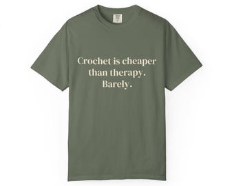 Puede incluir: Una camiseta de manga corta de color verde oliva con el texto "Crochet is cheaper than therapy. Barely." impreso en color crema claro en el centro del pecho. La prenda tiene un cuello redondo y un diseño de corte estándar.
