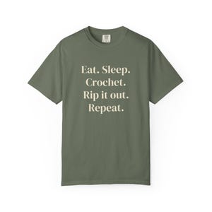 May include: An olive green short-sleeve t-shirt featuring the cream-colored text "Eat. Sleep. Crochet. Rip it out. Repeat." printed in a centered serif font on the front. The shirt has a crew neckline and a standard fit.