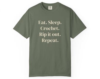 Puede incluir: Una camiseta de manga corta de color verde oliva con el texto en color crema "Eat. Sleep. Crochet. Rip it out. Repeat." impreso en una fuente serif centrada en la parte delantera. La camiseta tiene cuello redondo.