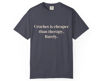 May include: A dark charcoal grey short-sleeve t-shirt featuring the cream-coloured text "Crochet is cheaper than therapy. Barely." printed in a serif font across the centre of the chest. The shirt has a crew neckline and a standard fit.
