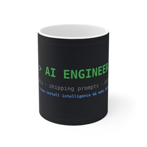 Puede incluir: Una taza de cerámica negra con interior blanco que presenta texto verde que dice AI ENGINEER, seguido de texto gris que dice shipping prompts, y texto azul que dice brew install intelligence && make deploy.