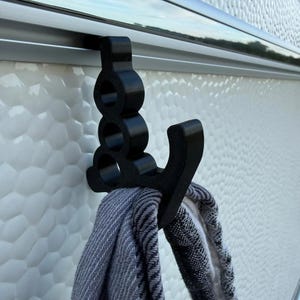 May include: A black plastic hook with a vertical design featuring three circular cutouts, mounted onto a horizontal metal rail. The hook has a curved arm extending outward, currently holding a folded grey and white patterned fabric item.