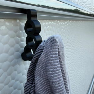 May include: A black, three-ringed hook designed to slide onto a horizontal rail. The hook is constructed with a vertical series of three circular openings and is currently holding a folded grey and white herringbone patterned fabric towel.