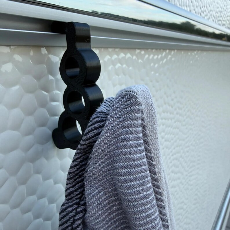 May include: A black, three-ringed hook designed to slide onto a horizontal rail. The hook is constructed with a vertical series of three circular openings and is currently holding a folded grey and white herringbone patterned fabric towel.