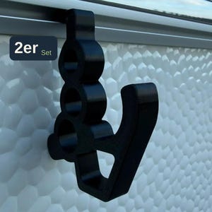 May include: A black plastic hook designed to hang over a ledge, featuring a vertical column with three circular cutouts and a curved arm extending outward. A small rectangular label in the upper left corner displays the text 2er Set.
