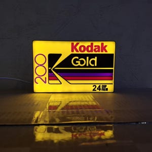 Może przedstawiać: Prostokątny podświetlany szyld z wzorem pudełka filmu Kodak Gold 200. Na żółtym tle znajduje się czerwone logo Kodak, słowo Gold, liczba 200 oraz napis 24 EXP FILM czarną czcionką, z poziomymi fioletowymi i czerwonymi paskami.