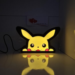 Puede incluir: Una lámpara de personaje iluminada en amarillo y negro con forma de criatura de dibujos animados con orejas largas, ojos redondos y manchas rojas en las mejillas. La lámpara tiene un borde negro, un cable de alimentación y un letrero Coup de coeur 78.