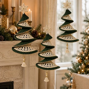 Puede incluir: Tres adornos navideños colgantes en forma de árbol en espiral, hechos de hilo verde oscuro y crema. Cada árbol presenta un copo de nieve de ganchillo en la parte superior, un diseño en espiral con cuentas doradas en los bordes y una borla crema.