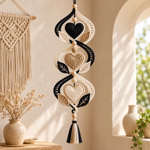 May include: A vertical macrame wall hanging featuring a spiral design with black, cream, and tan crocheted hearts and leaves. The piece is accented with round wooden beads and finished with a multi-colored tassel at the bottom end.