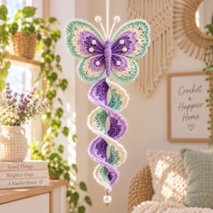 May include: A hanging crochet butterfly ornament featuring a purple, mint green, and cream color palette. The butterfly has a spiraling tail extending downward, finished with a single white pearl bead at the bottom, suspended by a white cord.