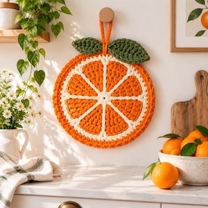 Pode incluir: Uma decoração de parede redonda em crochê com formato de fatia de laranja, apresentando segmentos de fio laranja brilhante separados por linhas brancas, rematada com duas folhas verdes em crochê e uma alça laranja para pendurar.