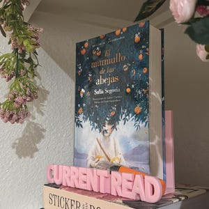 May include: A stack of three books with a pink bookend reading CURRENT READ. The top book is El Murmullo de las Abejas by Sofia Segovia with an orange tree cover. The middle book is Sticker Book II by John Derian. The bottom book is titled Woodlandia.