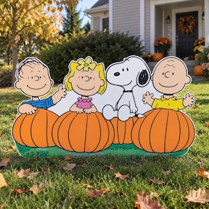 May include: A decorative yard sign featuring cartoon characters Linus, Sally, Snoopy, and Charlie Brown, each sitting inside a large orange pumpkin. The characters are illustrated with black outlines and vibrant colors, set against a green base.