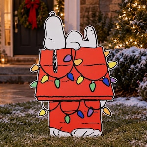 May include: A flat, outdoor garden decoration featuring the cartoon character Snoopy lying on top of a red doghouse. The doghouse is decorated with a string of colourful Christmas lights in yellow, green, blue, and purple, with black outlines.