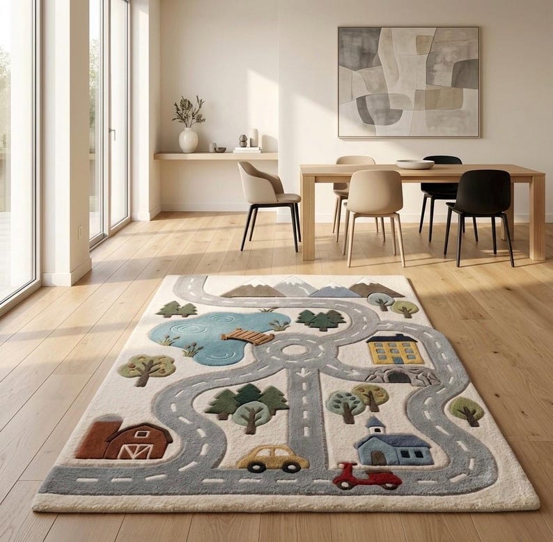 May include: A rectangular area rug featuring a play mat design with a light cream background. The rug displays a gray road network, a blue pond with a wooden bridge, green trees, brown mountains, a red barn, a yellow house, and a blue building.