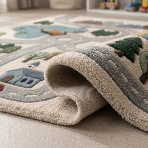 May include: A cream-colored area rug with a textured, tufted surface featuring a printed map design. The rug displays a winding gray road, blue houses, green trees, and a light blue lake. One corner of the rug is folded over, revealing the backing.