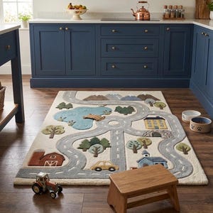 May include: A rectangular area rug featuring a tufted landscape design with a winding gray road, a blue pond with a wooden bridge, green trees, a red barn, a yellow house, a blue building, and a small yellow car on an off-white background.