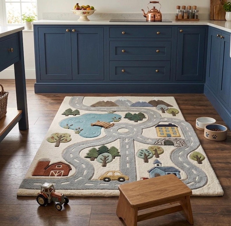 May include: A rectangular area rug featuring a tufted landscape design with a winding gray road, a blue pond with a wooden bridge, green trees, a red barn, a yellow house, a blue building, and a small yellow car on an off-white background.