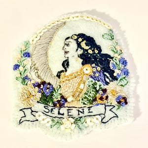 Puede incluir: Un parche bordado a mano sobre tela azul claro que muestra el perfil de una figura con cabello oscuro, una gran luna creciente y detalles florales. El diseño incluye hilo dorado, lentejuelas blancas y una cinta en la base con el texto SELENE.