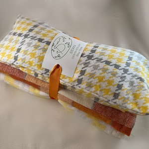May include: A stack of three fabric items tied with an orange ribbon. The top item has a yellow, gray, and white houndstooth pattern. The middle item has a yellow and orange plaid pattern. The bottom item has an orange and white plaid pattern. A tag reads "SEWN DESIGN ATELIER New York."