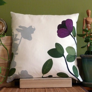 May include: Square throw pillow with a floral design. The pillow features a cream-colored background with a large purple flower and green leaves. A gray floral silhouette is also present. The pillow is placed on a wooden surface.
