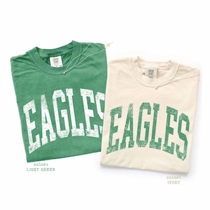 Könnte beinhalten: Zwei kurzärmelige T-Shirts mit Rundhalsausschnitt. Eines ist hellgrün mit weißem, abgenutztem Schriftzug EAGLES. Das andere ist elfenbeinfarben mit grünem, abgenutztem Schriftzug EAGLES. Beide Shirts haben ein Comfort Colors Nackenetikett.