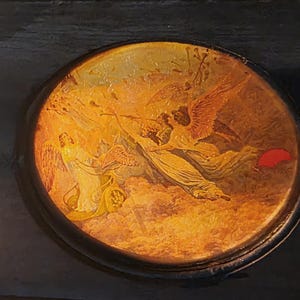 May include: A round, black-rimmed decorative box lid featuring a detailed, warm-toned illustration of winged figures in a chariot amidst clouds. The artwork displays golden and orange hues with a small, distinct red accent on the right side.