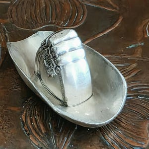 Könnte beinhalten: Eine silberfarbene Miniatur-Cowboyhut-Figur mit einer glatten, geschwungenen Krempe und einer strukturierten Krone. Die Seite der Krone zeigt ein detailliertes, geprägtes Blumenmuster mit komplexen Blütenblattstrukturen.
