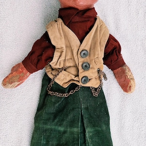 May include: A vintage doll with a painted red face, black mustache, and green cap. The doll wears a dark red shirt, a tan corduroy vest with three black buttons and a metal chain, and long green corduroy trousers. The hands are painted red.