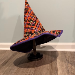 May include: A quilted witch hat featuring a vibrant orange, purple, and yellow plaid pattern. The hat has a tall, pointed crown and a wide brim with a purple fabric trim. A black ribbon with small orange pumpkin motifs encircles the base.