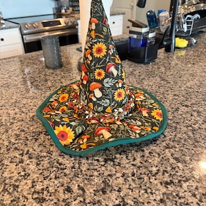 May include: A quilted fabric witch hat featuring a dark green background with a pattern of yellow sunflowers, orange and white mushrooms, and green foliage. The hat has a wide brim, a pointed crown, and a decorative ring of small twigs and berries.