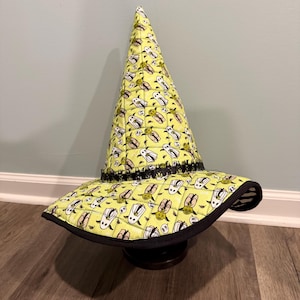 May include: A quilted, pointed hat in a light lime green color featuring a repeating pattern of small white ghost figures and green alien characters. The hat has a wide, slightly curved brim with a black fabric trim and a black decorative band.
